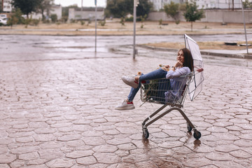 Beauty woman sitting in shopping cart with flowers and she hold in her hand umbrella. She is look happy and fun