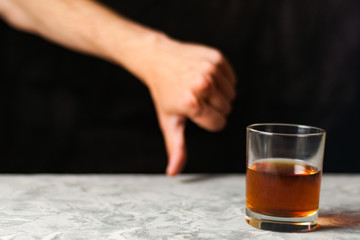 Man hand showing thumb down beside transparent glass with alcohol on gray cement on black background