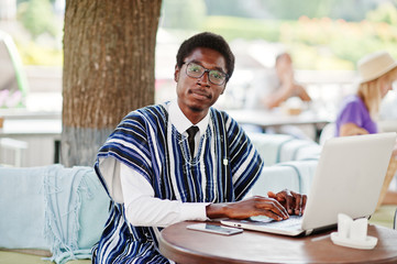 African man in traditional clothes and glasses sitting behind laptop at outdoor caffe and working.