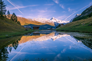 Snowy Mountains and Wood Chalet Reflecting in Altitude Lake at Sunset.
