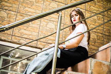 A fashionable and stylish girl in wide black pants, a white T-shirt with sunglasses with a white rim sits on the steps of the house outdoors and holds a jeans jacket in her hands