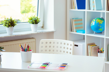 Interior of child room with white table and colorful pencils on him and shelf with books