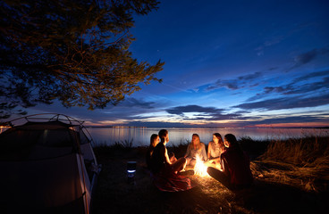 Night summer camping on sea shore. Group of five young tourists sitting on the beach around campfire near tent under beautiful blue evening sky. Tourism, friendship and beauty of nature concept. - obrazy, fototapety, plakaty
