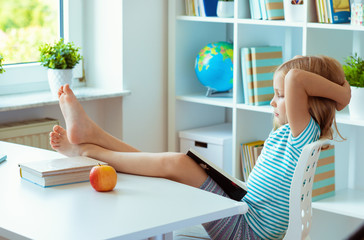 Portrait of funny little school girl relaxing reads book at the table in room at home