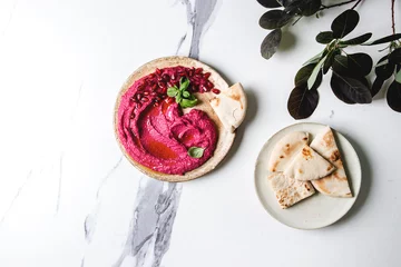 Schilderijen op glas Homemade spread beetroot hummus with pomegranate seeds, olive oil, basil served on ceramic plate with pita bread and green branch over white marble background. Flat lay, space. Mediterranean snack. © Natasha Breen