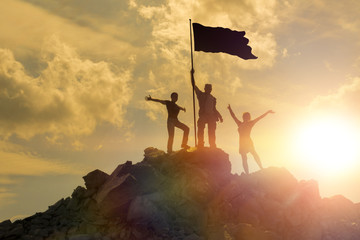 Silhouettes of happy three people on top of a mountain with the flag of victory. Success and...