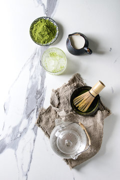 Ingredients for making matcha ice drink. Green tea matcha powder in ceramic bowl, traditional bamboo spoon, whisk, milk, glass teapot, ice cubes over white marble background. Flat lay, space