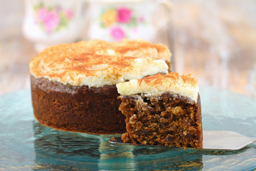 Slice of delicious walnut and carrot cake with marzepain icing with whole cake in the background
