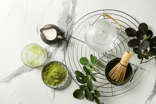 Ingredients for making matcha ice drink. Green tea matcha powder in ceramic bowl, traditional bamboo spoon, whisk on cooling rack, glass teapot, ice cubes over white marble background. Flat lay, space
