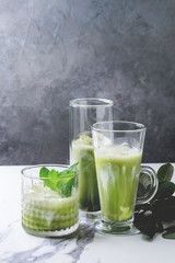 Matcha green tea iced latte or cocktail in three different glasses with ice cubes, matcha powder and jug of milk on white marble table, decorated by green branches. Grey wall at background