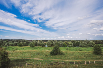 Fototapeta na wymiar Beautiful countryside landscape. Bright blue sky with charming white clouds, green trees, meadow, river, rural road and old wooden fence in village of Ukraine. Horizontal color photography.