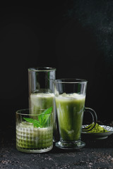 Matcha green tea iced latte or cocktail in three different glasses with mint, ice cubes and matcha powder over dark texture background