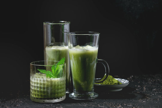 Matcha green tea iced latte or cocktail in three different glasses with mint, ice cubes and matcha powder over dark texture background