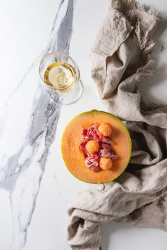 Melon and ham or prosciutto salad served in half of Cantaloupe melon on linen cloth over white marble background with glass of white wine. Flat lay, space