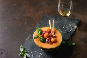 Melon and ham salad served in half of Cantaloupe melon with fresh basil and grissini bread on black slate board over dark brown texture background with glass of white wine.