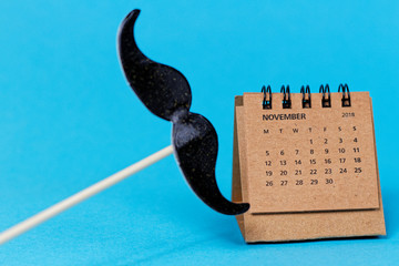 Fake mustache on the stick and paper calendar with November 2018 open page on blue background - symbol of Movember, event to raise awareness prostate and testicular cancer and men's suicide