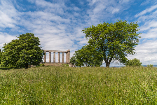 Edinburgh, Scotland, UK - June 13, 2012: National Monument is series of brown pilars against blue sky with white clouds. Green grass up front. Two trees.