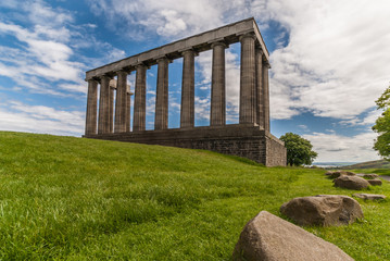 Fototapeta na wymiar Edinburgh, Scotland, UK - June 13, 2012: National Monument is series of dark brown pilars against blue sky with white clouds. Green grass up front with boulders.