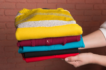 women's hands hold a stack of folded colored women's sweaters, against the backdrop of a terracotta brick wall, the concept of autumn and shopping