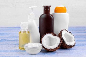 Various bottles of cosmetics with coconut extract next to fresh coconut on a blue wooden table on a white background