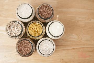 Various uncooked cereals, grains, and pasta for healthy cooking in glass jars on wooden table, white background, horizontal composition. Clean eating, balanced dieting food