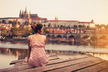 Young woman sitting on wood pier looking Prague Castle, Prague river Vltava and many famous Prague sights and historic architecture, back view. - obrazy, fototapety, plakaty