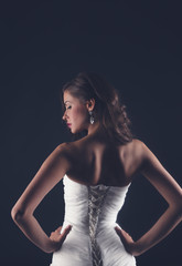 Beautiful slender young woman in white corset wedding or evening dress, stands with her back on a dark background, beauty and fashion, glamor