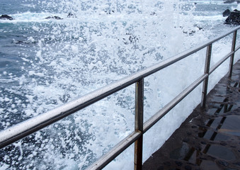 Dramatic white high foaming waves crashing over steel railings on a jetty a the edge of a summer blue sea