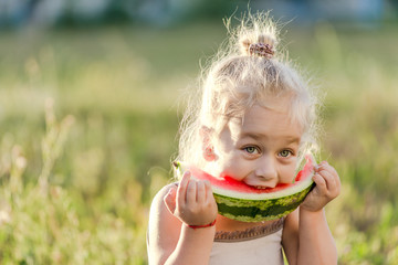 Little blond girl eating watermelon in the park.