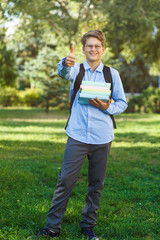 cute, young boy in round glasses in blue shirt with backpack holds books and thumb up. Education, back to school concept