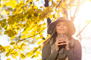 Young woman enjoying coffee outdoors on sunny autumn day. Closeup of female model standing by the tree with yellow leaves, with eyes closed holding coffee mug. Natural lighting, no retouch.