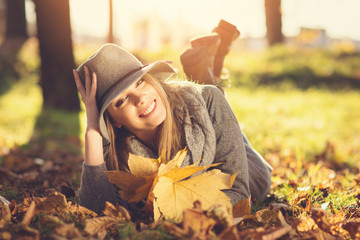 Cheerful woman enjoying nature outdoors in park, lying down on the grass and leaves on sunny autumn day. Natural lighting, mild retouch, filter applied.