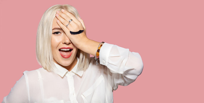 Young happy woman with eyelashes painted on the hand. Cute blonde hair model in white shirt closed her eye with hand. Concept of fun and merriment. Studio portrait on pink background.