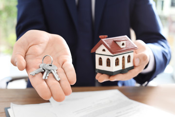 Real estate agent holding house model and keys at table in office, closeup