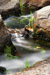 Detail view of flowing water of a small river, the water flows between large stones that are partially covered with moss and grass, long time exposure, flow structure, colors in the water