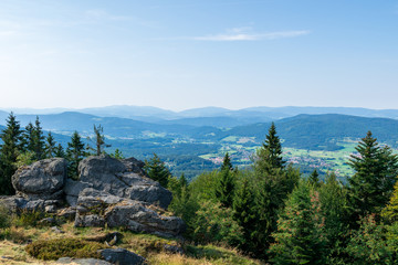 View from top of a mountain in the valley with clouds on the sky and mountains on the background and stones and trees in front of in the bavarian forest