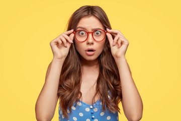 Shocked attractive teenager wears glasses with red rim, polka dot blouse, stares at camera with frightened expression as hears horrified news, poses against yellow background. Omg, its dreadful!