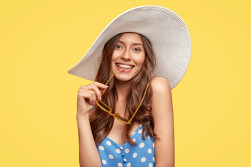 Good looking comely merry woman wears stylish summer hat, blue polka dot clothes, holds shades, being impressed by unforgettable vacation, poses indoor against yellow wall. People, holidays concept