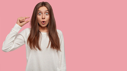 Surprised emotive young female with stupefied expression, gestures indoor, shows something very tiny, dressed in white sweater, stands against pink wall with copy space for your advertisement