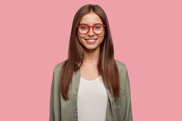 Studio shot of attractive student has gleeful expression, toothy smile, being in high spirit after lectures, dressed casually, wears round spectacles stands against pink background. Lively girl indoor