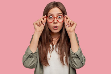 Studio shot of astonished shocked woman stares through spectacles, wears casual shirt, listens her...
