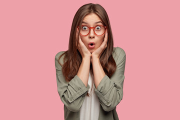 Terrific young female stares with bugged eyes, keeps hands on both cheeks, recieves bad news from interlocutor, wears round spectacles, isolated over pink background. People, amazement, shock