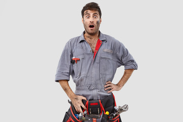 Disturbed surprised craftsman wears tool belt and overalls, reacts on sudden work, keeps hand on...