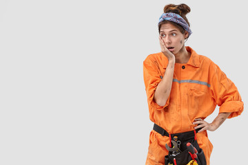 Indoor shot of desperate stunned untidy woman technician keeps hand on cheek, regrets something, has concentrated gaze aside, feels tired after hard work on factory, isolated over white wall