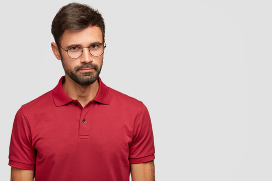 Attractive self confident hipster looks directly at camera with serious expression, wears round spectacles and red t shirt, stands against white background with copy space for your slogan. Masculinity