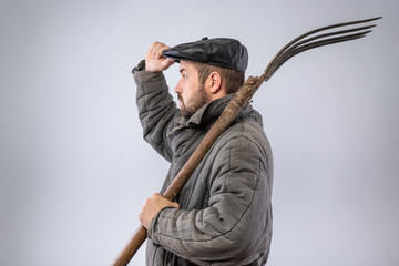 Man with pitchfork on his shoulder gives greeting gesture, dressed in old-fashioned clothes - cap and wool-padded jacket - Telogreika, studio photo. Idea - life in the village