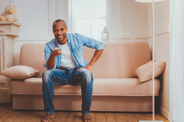 My day off. Content afro-american man sitting on the sofa and holding a remote control