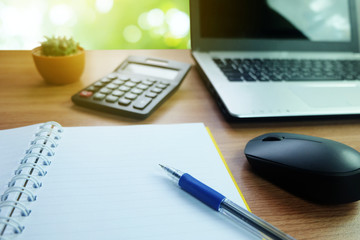 Notebook with empty space and a pen and calculator ,with laptop computer and wireless mouse on the desk wooden with cactus and Bokeh background