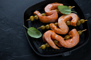 Cast-iron grill with raw marinated pork loin and green olives on skewers, studio shot over black stone surface