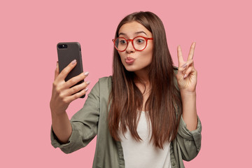 Pleasant looking young female pouts lips, makes grimace, shows peace sign at camera of modern smart phone, poses for selfie, shares photos with friends, makes video call. Youth and technology